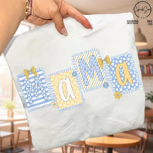 May include: White t-shirt featuring the word "Mama" in a patchwork style. The letters are in blue and yellow, with floral and striped patterns. The design includes bows and small flowers.