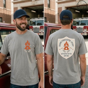 May include: A grey t-shirt featuring a small orange flame and axe graphic on the front left chest. The back displays a large shield-shaped emblem with the text DOING FIREFIGHTER STUFF AND PRAYING THE TONS DON'T DROP, centered above a flame and axe icon.