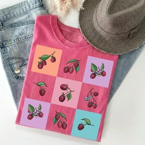 Könnte beinhalten: Ein pinkfarbenes T-Shirt mit einem Rastermuster aus Himbeer-Illustrationen auf farbigen Quadraten. Das Shirt wird auf einer blauen Jeans präsentiert, mit einem Fedora-Hut und einer Halskette im Hintergrund.
