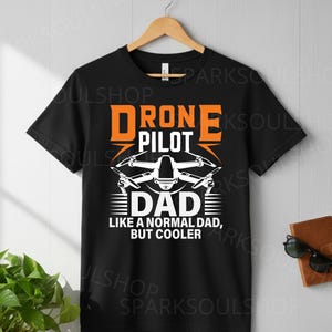 May include: Black t-shirt with the words "DRONE PILOT DAD" in orange and white. The design includes a drone graphic and the phrase "LIKE A NORMAL DAD, BUT COOLER". The shirt is hanging on a wooden hanger.