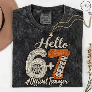 May include: A charcoal grey acid-wash t-shirt featuring the text Hello 6+7, the word SIX inside the number 6, the word SEVEN inside the number 7, and the text #OfficialTeenager. The shirt has a Comfort Colors label at the inner neckline.