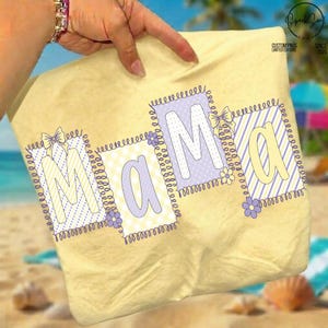 May include: A light yellow beach towel featuring the word "MAMA" in pastel colours. Each letter is in a square with a different pattern, including polka dots and stripes, and is decorated with bows and flowers. The towel is held by a hand.