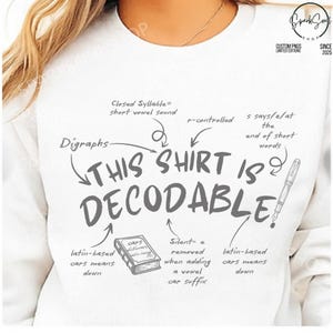 Op de afbeelding: Een wit sweatshirt met ronde hals en de donkergrijze tekst THIS SHIRT IS DECODABLE!, omringd door educatieve taalkundige annotaties zoals Digraphs, Closed Syllable, r-controlled en s says /z/ at the end of short words, inclusief een boek en pen.