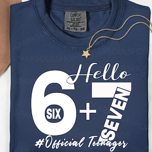 May include: Navy blue t-shirt with the text "Hello 6 + 7 Seven #Official Teenager" in white. A gold-colored necklace with a star pendant is draped on the shirt. The shirt has a Comfort Colors tag.