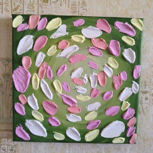 May include: Square canvas art with a textured, abstract floral design. The artwork features a green background with a central lighter green circle. Pink, white, and yellow brushstrokes create a petal-like pattern, adding depth and dimension.