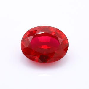 May include: A vibrant, oval-cut gemstone with a deep, saturated red color. The stone features a faceted surface that reflects light, creating a brilliant, sparkling appearance across its polished, symmetrical, and translucent red surface.