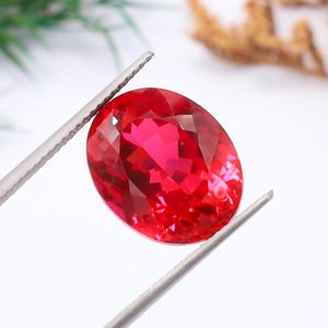 May include: A vibrant, oval-cut red gemstone held by silver-toned metal tweezers. The stone features a faceted surface that reflects light, creating deep crimson highlights and shadows throughout its transparent, polished crystalline structure.