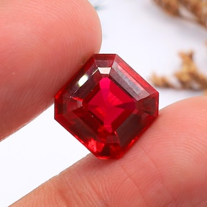 May include: A faceted, square-cut, bright red gemstone. The gem is held between fingers, showcasing its vibrant color and geometric precision. The gem's facets reflect light, creating a sparkling effect. This gemstone is likely for jewelry making.