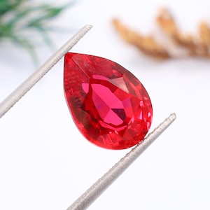 Puede incluir: Una gema roja vibrante en forma de pera sostenida por unas pinzas de metal plateado. La piedra facetada muestra un tono carmesí brillante y translúcido con cortes geométricos nítidos que reflejan la luz en su superficie pulida.