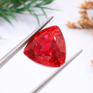 Puede incluir: Una gema roja brillante, facetada y de forma triangular, es sostenida por unas pinzas plateadas. La gema tiene muchas superficies reflectantes, que capturan la luz. El fondo está borroso, con toques de verde y marrón.