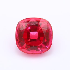 Puede incluir: Una gema de rubí de corte cojín vibrante con un color rojo profundo y saturado. La piedra presenta una superficie facetada que crea un patrón simétrico de reflejos claros y oscuros, destacando su calidad transparente y acabado pulido.