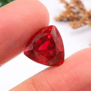 Peut inclure: Une pierre précieuse rouge vif et profonde taillée en forme triangulaire avec de multiples facettes qui reflètent la lumière. La pierre présente une clarté translucide brillante et une forme géométrique nette, affichant une riche teinte cramoisie.