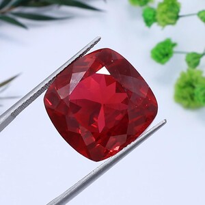 May include: A vibrant, cushion-cut red gemstone held by silver-toned metal tweezers. The stone features a faceted surface that reflects light, showcasing a deep, saturated crimson hue throughout its transparent, polished structure.