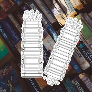 May include: Two white stickers shaped like stacks of books, topped with floral designs. The stickers have a reading tracker design with the text "Reading Tracker 2026". The stickers are set against a backdrop of old books.