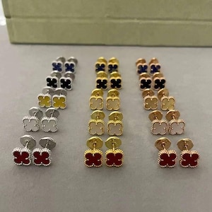 Op de afbeelding: Een verzameling klavervormige oorstekers in verschillende kleuren, waaronder rood, wit, geel, zwart en blauw. De oorbellen hebben zilver- en goudkleurige metalen pinnen.