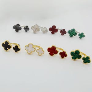 Puede incluir: Una colección de anillos con forma de trébol en varios colores, incluyendo negro, blanco, rojo y verde. Los anillos son de metal y presentan un diseño de doble trébol. Algunos anillos tienen una banda dorada, otros una banda plateada.