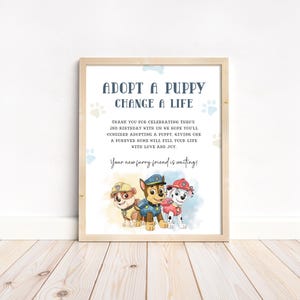 May include: A framed print with the text "ADOPT A PUPPY CHANGE A LIFE" and a message about adopting a puppy. The print features cartoon puppies in various colors. The frame is light brown.