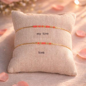 Peut inclure: Deux bracelets dorés avec des perles corail reposent sur un petit coussin beige. Le bracelet supérieur indique "my love" et le bracelet inférieur "love". Le fond présente des pétales de roses roses et un éclairage doux.