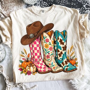 Puede incluir: Una camiseta color crema con un gráfico de un sombrero de vaquero marrón sobre tres botas vaqueras. Las botas tienen patrones de cuadros rosas, flores y manchas de vaca. El diseño incluye calabazas naranjas, tallos de trigo y flores.