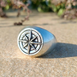 Könnte beinhalten: Ein silberner Ring mit einem Kompassrosen-Design. Der Ring hat eine abgerundete Form und eine polierte Oberfläche. Die Kompassrose hat acht Punkte in einem kreisförmigen Rahmen. Der Ring liegt auf einer strukturierten Oberfläche.