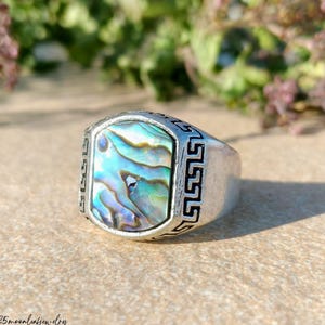 May include: A silver ring with a rectangular abalone shell inlay. The iridescent shell features shades of blue, green, and purple. The ring has a Greek key design around the shell and on the sides. The shop name "125moonleafjewelry" is at the bottom.