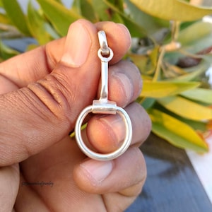 May include: A silver-colored pendant with a circular ring at the bottom and a loop at the top. The pendant is held in a hand against a background of green leaves.