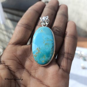 Op de afbeelding: Een ovale turkoois hanger met een zilveren zetting. De steen is helderblauw met bruine en gouden aders. De hanger wordt in een hand gehouden. De tekst "925moonleafjewelry" is zichtbaar.