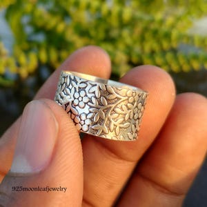 Op de afbeelding: Een zilveren ring met een bloemmotief. De brede ring heeft een gedetailleerd patroon van bladeren en kleine bloemen. De ring wordt in een hand gehouden, met de tekst "925moonleafjewelry" zichtbaar.