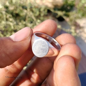 May include: A silver signet ring with a smooth, oval face. The ring is held between fingers, showcasing its polished metal finish. The ring's design is simple and classic, suitable for everyday wear. The text "125moonleafjewelry" is visible in the bottom left corner.
