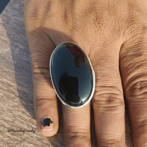 Può includere: Un anello con una grande pietra nera ovale e una fascia argentata. La pietra nera lucida riflette la luce, creando un aspetto lucido. L'anello è un gioiello importante, adatto a varie occasioni. Il testo "925moonleafjewelry" è visibile.