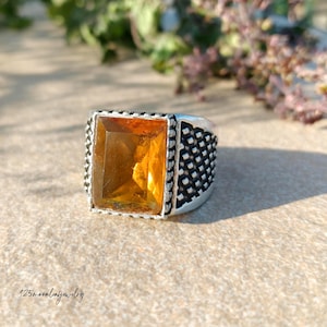 May include: Silver ring with a large, rectangular, amber-colored gemstone. The ring has a textured band and a decorative setting around the stone. The ring is displayed on a neutral surface with blurred greenery in the background.