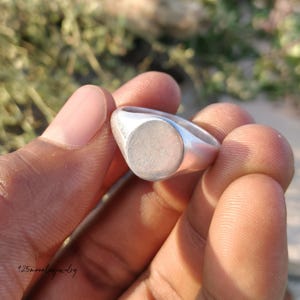 May include: A silver signet ring with a smooth, oval face. The ring is held in a hand against a blurred green and brown background. The ring's band is rounded and polished. The ring is a simple, classic piece of jewellery.