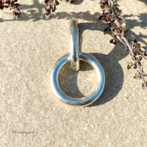 May include: A silver-colored ring pendant with a circular design and a connecting loop. The jewelry piece is displayed on a light brown surface, with dried plant stems in the background. The shop's name, "125moonleafjewelry", is written in cursive at the bottom.