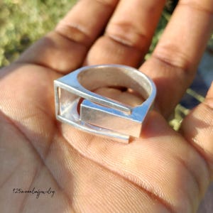 May include: A silver ring with a geometric design. The ring features a rectangular bar set within a square frame, creating an open, modern look. The ring is held in a hand, with the text "125moonleafjewelry" visible.