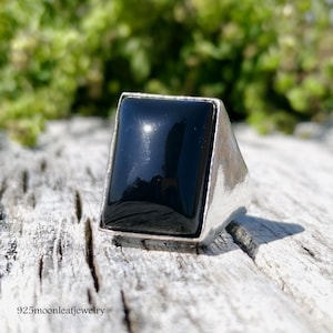 Może przedstawiać: Srebrny pierścionek z dużym, prostokątnym czarnym onyksem. Pierścionek ma polerowaną srebrną obręcz i oprawę w ramce. Tło to rozmyty widok zielonych liści. Tekst "925moonleafjewelry" znajduje się na dole.