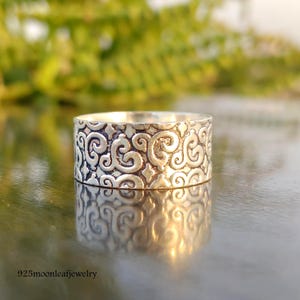 May include: A silver ring with an ornate, swirling design. The ring is wide and has a textured surface, reflecting light. The pattern is consistent around the entire band. The ring is displayed on a reflective surface.