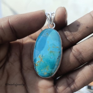 Op de afbeelding: Ovale turkoois hanger met een zilverkleurige balein. De turkoois steen heeft een levendige blauwe kleur met bruine en bruingele adering. De hanger wordt in een hand gehouden, wat het ontwerp en de kleur van het sieraad laat zien.