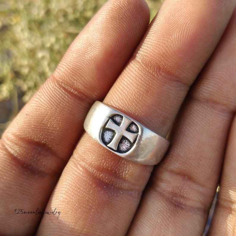Pu&ograve; includere: Un anello d'argento con un disegno a croce in rilievo. L'anello ha una fascia larga e un emblema a croce dettagliato. L'anello &egrave; mostrato su un dito, con il disegno a croce centrato sulla parte superiore. L'anello &egrave; probabilmente in argento sterling.