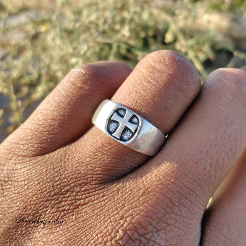 Pu&ograve; includere: Un anello d'argento con un motivo a croce. L'anello &egrave; largo e ha una finitura liscia e lucida. La croce &egrave; centrata sull'anello e ha uno sfondo strutturato e scuro. L'anello &egrave; su un dito.