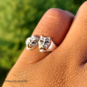 Puede incluir: Un anillo de plata con dos máscaras teatrales, una sonriente y otra fruncida. El anillo está diseñado con detalles intrincados, mostrando los símbolos clásicos de la comedia y la tragedia. El anillo se muestra en un dedo sobre un fondo verde borroso.