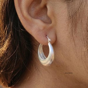 May include: A silver hoop earring is shown close up. The earring has a thick, rounded design and a smooth, matte surface. It is attached to an ear. The background is blurred, with glimpses of skin and hair.