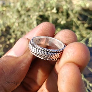 Puede incluir: Un anillo de plata con un diseño texturizado. El anillo presenta un patrón trenzado alrededor de los bordes y un patrón de espiga en el centro. El anillo se sostiene en una mano sobre un fondo verde borroso.