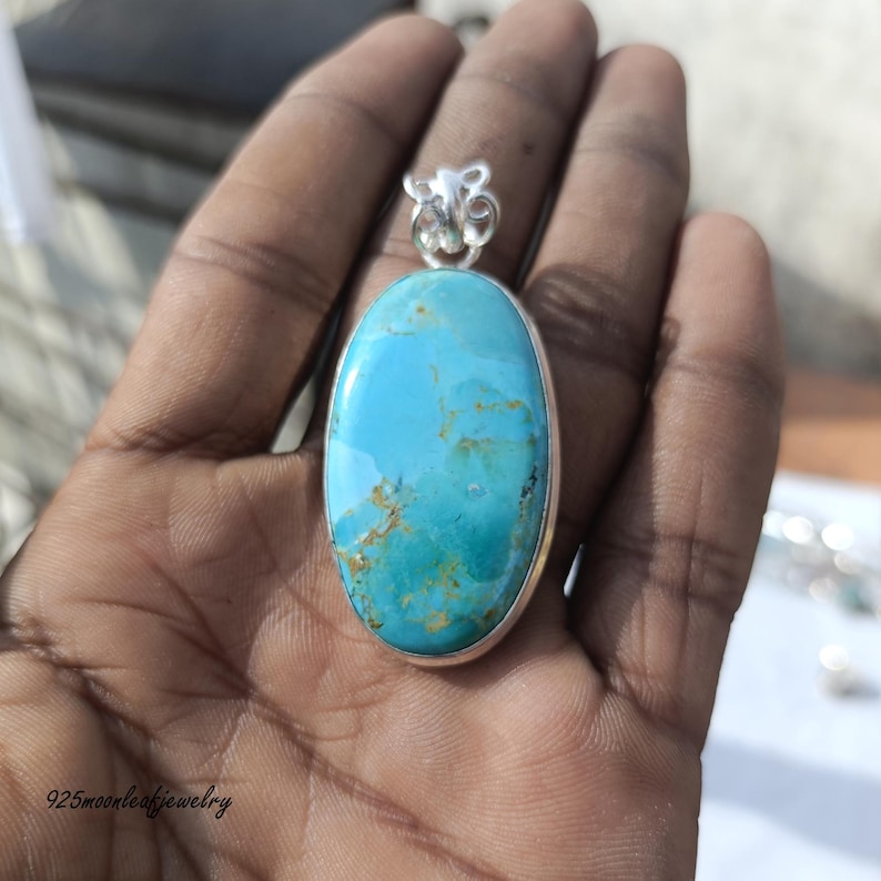 Op de afbeelding: Een turquoise hanger met een zilveren zetting. De ovaalvormige steen heeft een levendige blauwe kleur met bruine en gouden adering. De hanger wordt in een hand gehouden en de tekst "925moonleafjewelry" is onderaan zichtbaar.