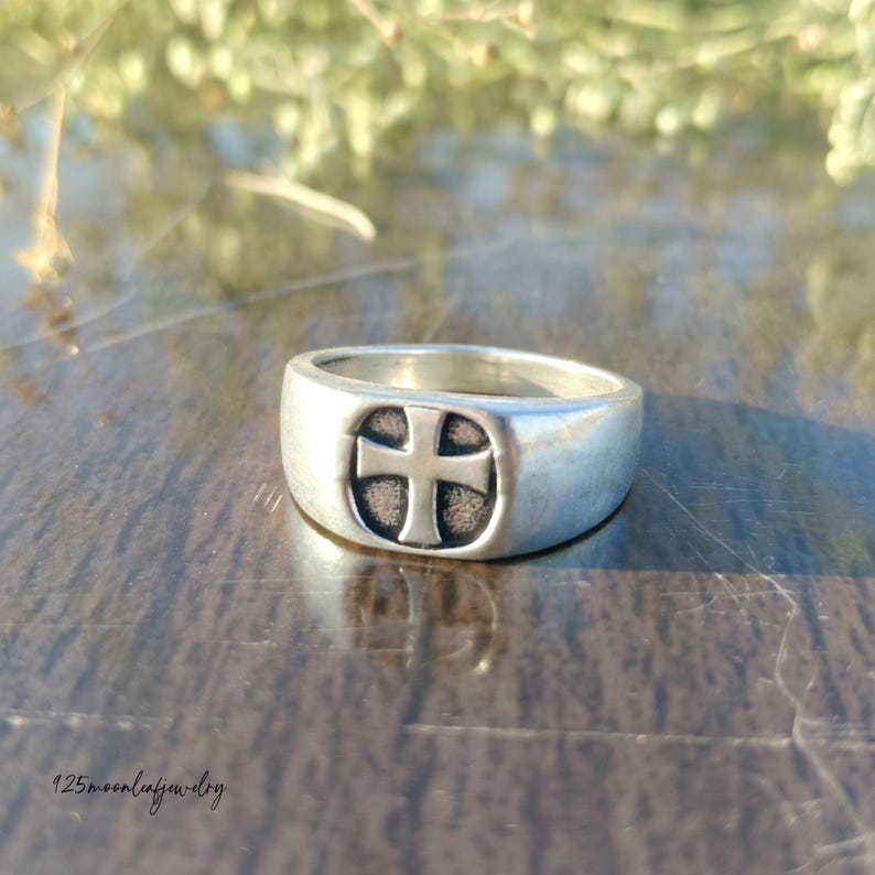 Pu&ograve; includere: Un anello d'argento con un design a croce. L'anello ha una fascia larga e una faccia ovale in rilievo con una croce al centro. L'anello &egrave; realizzato in un materiale metallico lucido.