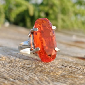 May include: A silver ring featuring a large, faceted, orange gemstone. The gemstone is an elongated oval shape, with a bright, fiery color. The ring is set on a wooden surface with the text "925moonleafjewelry".