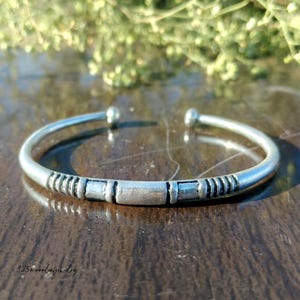 May include: A silver cuff bracelet with a tribal design. The bracelet has a central rectangular section flanked by shorter rectangular and striped sections. The ends of the bracelet have small, rounded ball-shaped accents. The bracelet is displayed on a brown surface.