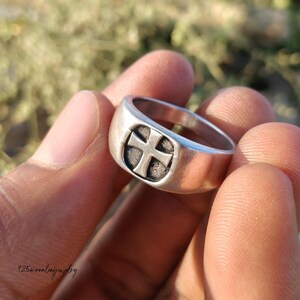 Pu&ograve; includere: Un anello d'argento con un motivo a croce. L'anello ha una fascia spessa e una faccia quadrata con una croce in rilievo. L'anello &egrave; tenuto in mano, con uno sfondo sfocato.