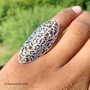 Könnte beinhalten: Ein silberfarbener Filigranring mit einer länglichen ovalen Form. Der Ring hat ein offenes Design mit komplizierten Wirbelmustern. Der Ring wird an einem Finger getragen, mit dem Text "925moonleafjewelry" unten sichtbar.