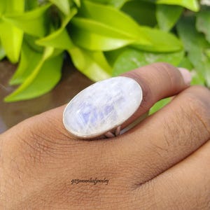 May include: A ring featuring a large, oval-shaped, translucent moonstone with a blue iridescent sheen, set in a simple silver-toned metal band. The stone displays a smooth, polished surface with visible internal light reflections and subtle blue hues.