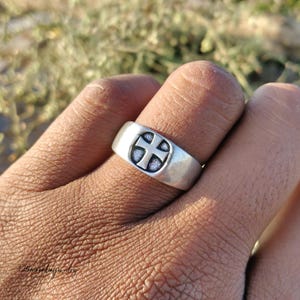 Pu&ograve; includere: Un anello d'argento con un motivo a croce. L'anello &egrave; largo e ha una finitura liscia e lucida. La croce &egrave; centrata sull'anello e ha uno sfondo strutturato e scuro. L'anello &egrave; su un dito.
